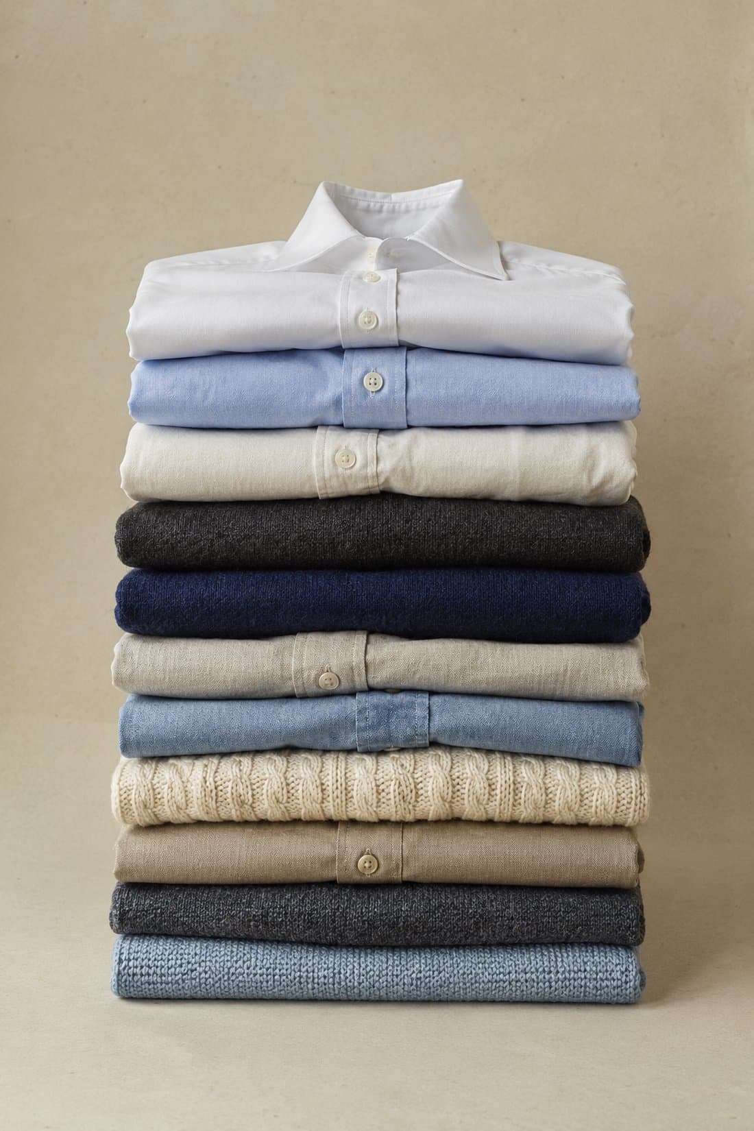 A stack of eleven folded shirts and knitwear in white, blue, stone and charcoal, resting on bone paper.